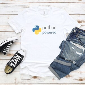 Peut inclure: T-shirt blanc avec un logo Python jaune et bleu et le texte "python powered". Le t-shirt est pos&eacute; &agrave; plat sur une surface en bois blanc avec une paire de baskets Converse noires et un jean bleu.