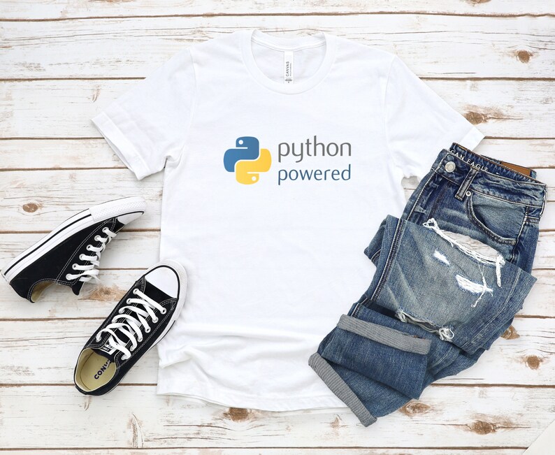 Peut inclure: T-shirt blanc avec un logo Python jaune et bleu et le texte "python powered". Le t-shirt est pos&eacute; &agrave; plat sur une surface en bois blanc avec une paire de baskets Converse noires et un jean bleu.