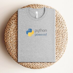 Peut inclure: Un t-shirt gris avec un logo Python bleu et jaune et le texte "python powered".