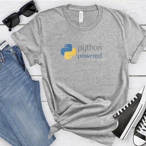 Peut inclure: Un t-shirt gris avec un logo Python bleu et jaune et le texte "python powered". Le t-shirt est pos&eacute; sur une surface en bois blanc avec des lunettes de soleil noires, un jean bleu et des baskets noires.