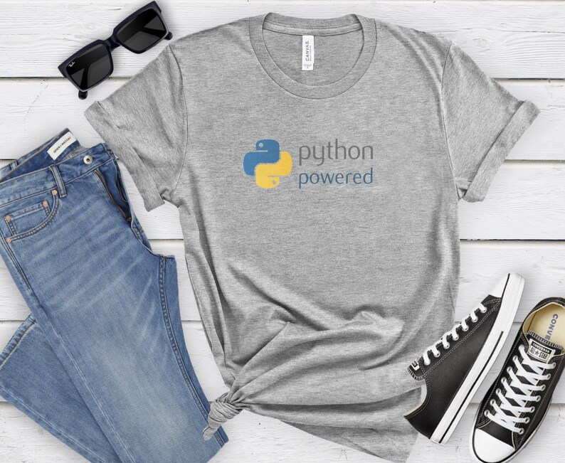 Peut inclure: Un t-shirt gris avec un logo Python bleu et jaune et le texte "python powered". Le t-shirt est pos&eacute; sur une surface en bois blanc avec des lunettes de soleil noires, un jean bleu et des baskets noires.
