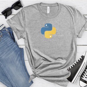 May include: A gray t-shirt with a yellow and blue Python logo on the front. The shirt is laid out on a white wooden surface with a pair of blue jeans, sunglasses, and black sneakers.