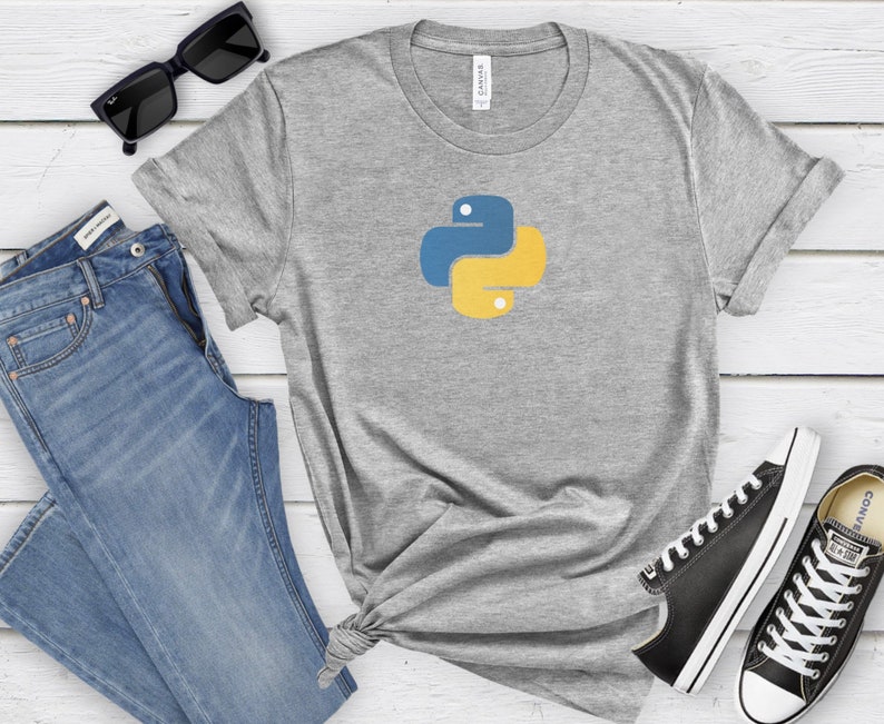 May include: A gray t-shirt with a yellow and blue Python logo on the front. The shirt is laid out on a white wooden surface with a pair of blue jeans, sunglasses, and black sneakers.