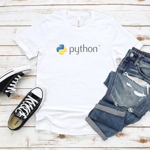 Peut inclure: T-shirt blanc avec un logo Python jaune et bleu et le mot "python" en texte noir. Le t-shirt est pos&eacute; sur une surface en bois blanc avec une paire de chaussures Converse noires et un jean bleu.
