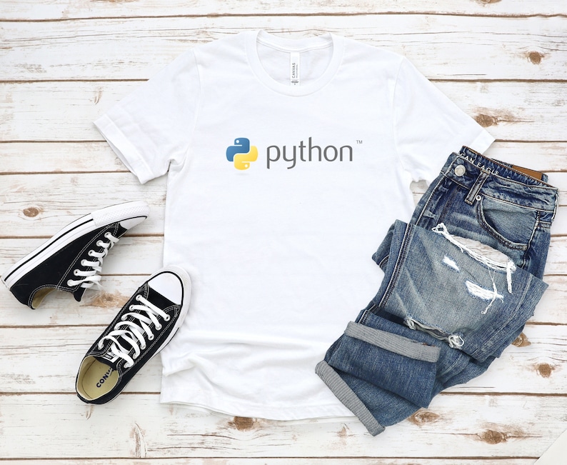 Peut inclure: T-shirt blanc avec un logo Python jaune et bleu et le mot "python" en texte noir. Le t-shirt est pos&eacute; sur une surface en bois blanc avec une paire de chaussures Converse noires et un jean bleu.