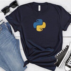 May include: A navy blue t-shirt with a yellow and blue Python logo. The t-shirt is on a white wooden surface with a pair of blue jeans, sunglasses, and black sneakers.