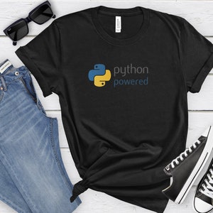 Peut inclure: T-shirt noir avec un logo Python jaune et bleu et le texte "python powered". Le t-shirt est pos&eacute; sur une surface en bois blanc avec des lunettes de soleil, un jean bleu et des baskets noires.