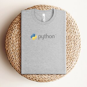 Peut inclure: Un t-shirt gris avec un logo Python jaune et bleu et le mot "python" en blanc.