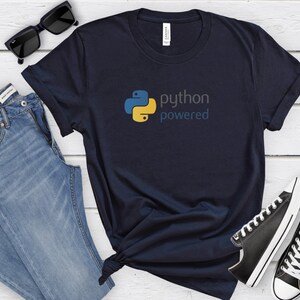 Peut inclure: Un t-shirt bleu marine avec un logo Python jaune et bleu et le texte "python powered". Le t-shirt est pr&eacute;sent&eacute; sur un fond en bois blanc avec des lunettes de soleil, un jean bleu et des baskets noires.