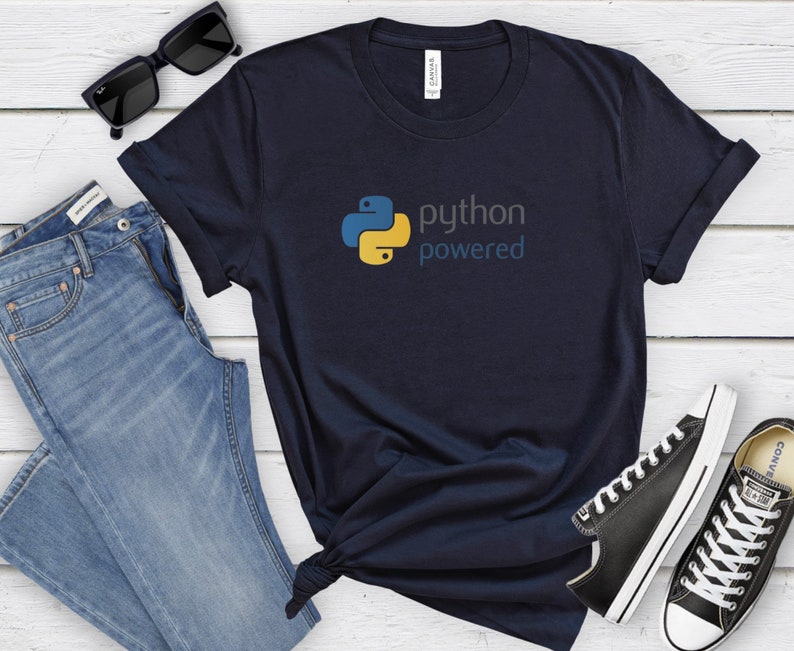 Peut inclure: Un t-shirt bleu marine avec un logo Python jaune et bleu et le texte "python powered". Le t-shirt est pr&eacute;sent&eacute; sur un fond en bois blanc avec des lunettes de soleil, un jean bleu et des baskets noires.