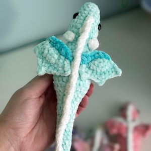 May include: A handmade, plush dragon toy in shades of light blue and white. The dragon features a textured, velvety material, with white accents along its spine and wings. The wings have a darker blue trim. The dragon has black eyes.