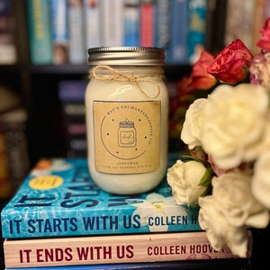 Op de afbeelding: Een witte kaars in een glazen pot met een bruin etiket waarop "Ray's Enchanted Candle Co." en "Cinnabon 100% All Natural Soy Wax" staat. De kaars staat op een stapel van drie boeken: "It Starts With Us", "It Ends With Us" en "Ugly Love", allemaal van Colleen Hoover.