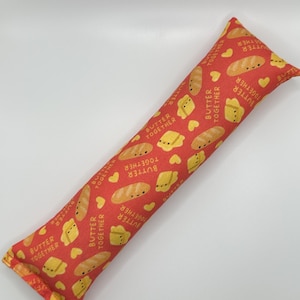 May include: A red and yellow cat toy shaped like a long pillow with a repeating pattern of butter and bread. The pattern includes the text "Butter Together".