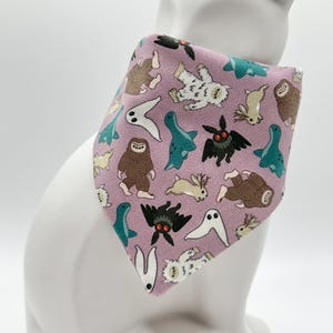 May include: A lavender pet bandana featuring a repeating pattern of cartoon cryptids, including a mothman, bigfoot, and other mythical creatures. The bandana is triangular and is draped around a white ceramic cat statue.