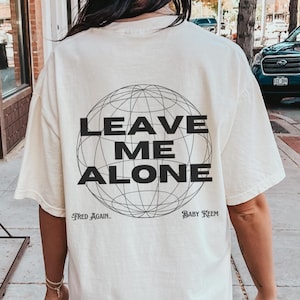 May include: A white t-shirt with a black graphic of a globe with the text "LEAVE ME ALONE" inside. The text "FRED AGAIN.." and "BABY KEEM" are below the globe.