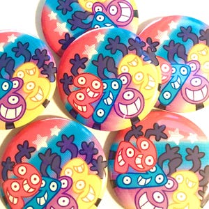 Puede incluir: Seis botones redondos coloridos con un fondo degradado de arcoíris y estrellas blancas. Cada botón presenta un personaje de dibujos animados con múltiples ojos y una amplia sonrisa. Los personajes son de varios colores, incluyendo rojo, amarillo, azul y morado.