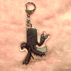 Könnte beinhalten: Ein schwarzer und grauer Buchcharakter-Schlüsselanhänger mit einem lila Augenklappen, der einen Stock hält und mit einer Hand winkt. Der Charakter hat ein zahniges Grinsen und sitzt auf einem grauen, pelzigen Schwanz.