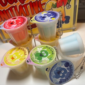 Peut inclure: Plusieurs petits gobelets en plastique avec des couvercles, chacun étiqueté avec une saveur différente : fraise, banane, matcha et myrtille. Les couvercles présentent des personnages de dessins animés et le texte "NOT REAL FOOD DO NOT EAT". Chaque tasse a une petite chaîne attachée.