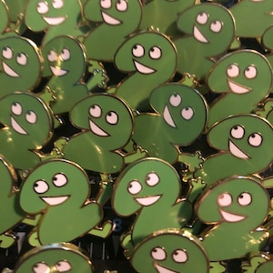 May include: A close-up of a group of green enamel pins shaped like the number two with a smiling face and three eyes. The pins have a gold outline and are arranged in a random pattern.