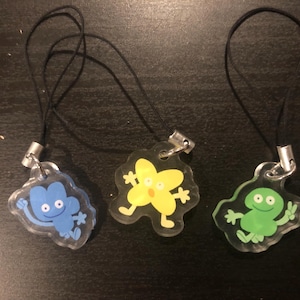 Puede incluir: Tres abalorios de personajes coloridos en cordones de teléfono negros. Los abalorios son azules, amarillos y verdes. El abalorio azul es un personaje de cuatro brazos con una expresión de sorpresa. El abalorio amarillo es una estrella de cinco puntas con una expresión de sorpresa. El abalorio verde es una rana con una expresión feliz.