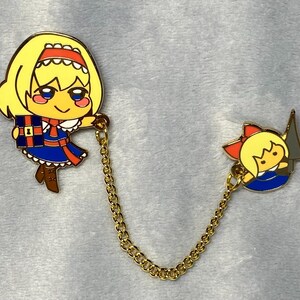 May include: Two gold-colored enamel pins connected by a gold chain. The first pin features a blonde-haired girl in a blue dress holding a blue and red gift box. The second pin features a blonde-haired girl in a blue dress holding a gray sword.