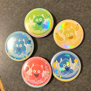 Könnte beinhalten: Fünf bunte, runde Buttons mit Cartoon-Charakter-Designs. Jeder Button hat ein anderes Farbschema: Grün, Gelb, Blau, Rot und Blau. Die Designs umfassen verschiedene Formen und Muster mit einem holografischen Effekt.
