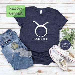 May include: A navy blue t-shirt with a white Taurus zodiac symbol and the word "Taurus" printed on the front. The shirt is made of a soft, comfortable material and is perfect for everyday wear.