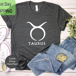 May include: Dark grey heather t-shirt with a white Taurus zodiac symbol and the word "TAURUS" printed on the front.