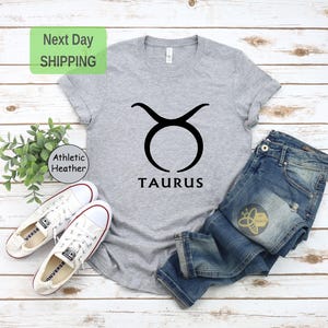 May include: Gray t-shirt with a black Taurus zodiac symbol and the word "Taurus" printed below it. The shirt is laid flat on a wooden surface with a pair of white sneakers and blue jeans.