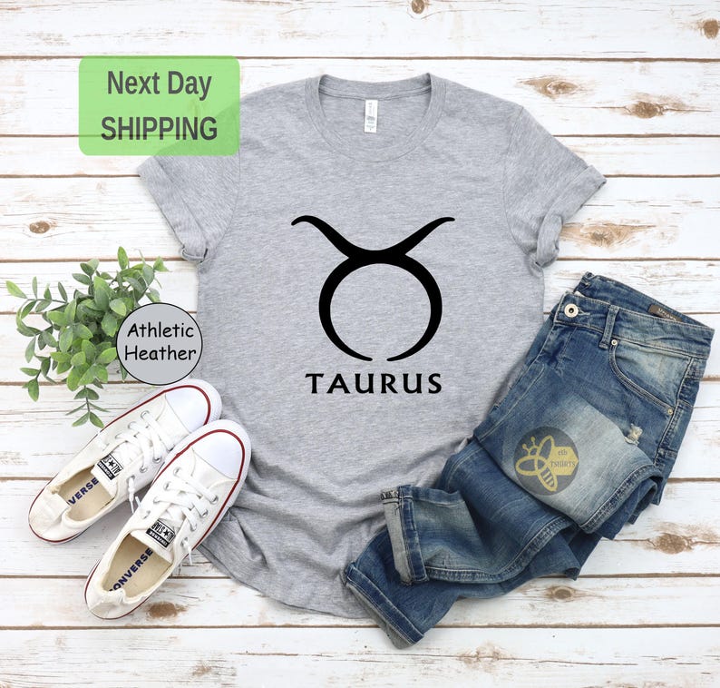 May include: Gray t-shirt with a black Taurus zodiac symbol and the word "Taurus" printed below it. The shirt is laid flat on a wooden surface with a pair of white sneakers and blue jeans.