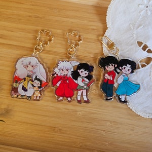 May include: Four colourful anime character keychains with gold metal rings. The keychains feature Inuyasha, Kagome, Sango, and Miroku from the anime series Inuyasha.