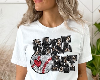 May include: White t-shirt with the words "GAME DAY" in a black and leopard print design. A baseball with a red heart is featured below the text. The shirt is paired with denim shorts.