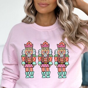 May include: A light pink sweatshirt featuring three colorful nutcracker illustrations. Each nutcracker has a pink and green crown, a white mustache, and is wearing a pink and green uniform with blue boots. The sweatshirt is a crew neck style.