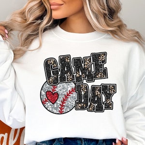May include: White sweatshirt with a black and white graphic print of a baseball with a red heart and the text "GAME DAY" in black with a leopard print pattern.