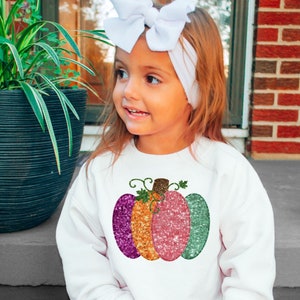 Puede incluir: Una sudadera blanca con un diseño de calabaza brillante en tonos de rosa, naranja y verde. La calabaza tiene un tallo marrón y hojas verdes.