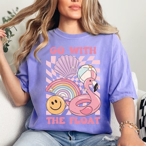 May include: A lilac-colored t-shirt with a graphic design featuring a pink flamingo float, rainbow, seashell, beach ball, and a smiley face. The text "GO WITH THE FLOAT" is printed in pink. The shirt is a relaxed fit.