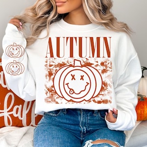 May include: White sweatshirt with the word "AUTUMN" in orange block letters and a pumpkin graphic with crossed-out eyes and a tongue sticking out. Two smaller pumpkin graphics are on the left sleeve.