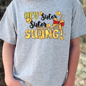 May include: A gray t-shirt with a graphic design featuring a baseball bat, baseballs, and the text "Hey Sister Sister Swing!"