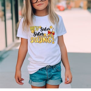 May include: White t-shirt with a graphic of a baseball bat and baseballs with the text "Hey Sister Sister Swing!"
