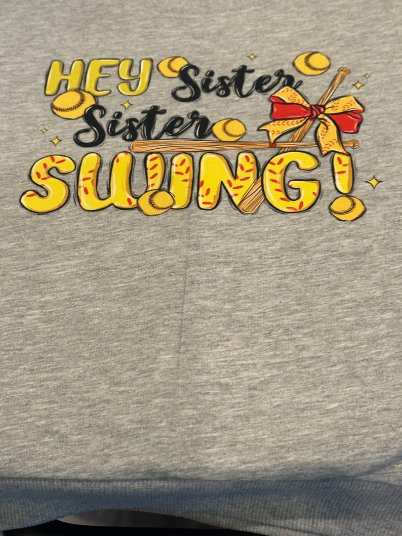May include: Gray t-shirt with a yellow and red graphic design that says "Hey Sister Sister Swing!" with baseball bats and baseballs.