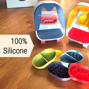 Könnte beinhalten: Tierfutterbehälter in verschiedenen Farben und Designs, darunter eine blaue Katzenschüssel und eine gelbe Hundeschüssel. Die Schalen bestehen aus 100 % Silikon und werden auf einem Holzboden präsentiert. Mehrere farbige Silikonschalen sind ebenfalls abgebildet.