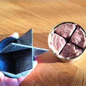 Puede incluir: Una herramienta negra y azul impresa en 3D con un divisor en forma de cruz, junto a una lata abierta de comida dividida en cuatro porciones iguales. La comida es de color rosa claro. Los artículos están sobre una superficie de madera.