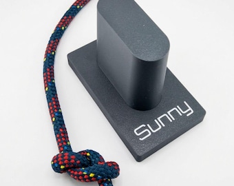 May include: A dark gray, rectangular base with a rounded, upright pillar and the word Sunny printed in white. A dark blue rope with red and yellow flecks is tied in a knot and curves around the base, featuring a textured, braided surface.