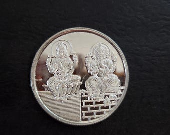 May include: A silver coin with a raised design of two Hindu deities, Lakshmi and Ganesha, sitting on a platform. The coin is circular and has a smooth, reflective surface.
