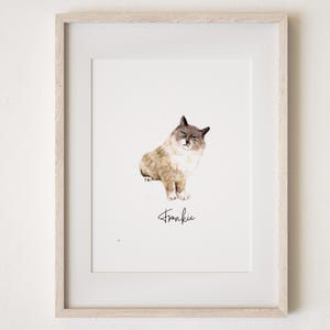 May include: A framed watercolour print featuring a cat named Frankie. The cat is rendered in brown, beige, and grey tones, with a white chest and paws. The cat is seated and facing forward. The name "Frankie" is written in cursive script beneath the cat.