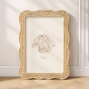 Könnte beinhalten: Ein gerahmter Druck mit einer Aquarellillustration eines flauschigen, hellen Hasen mit langen Ohren. Der Druck ist in einem natürlichen, gewebten Weidenrahmen mit gewelltem Rand eingefasst. Das Wort "Flopsy" ist darunter geschrieben.