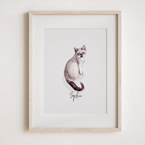 May include: Framed watercolour painting of a Siamese cat named Sophie. The cat is depicted in shades of grey, brown, and black, with blue eyes. The cat is sitting and looking towards the viewer. The name "Sophie" is written in cursive below the cat. The frame is light wood.