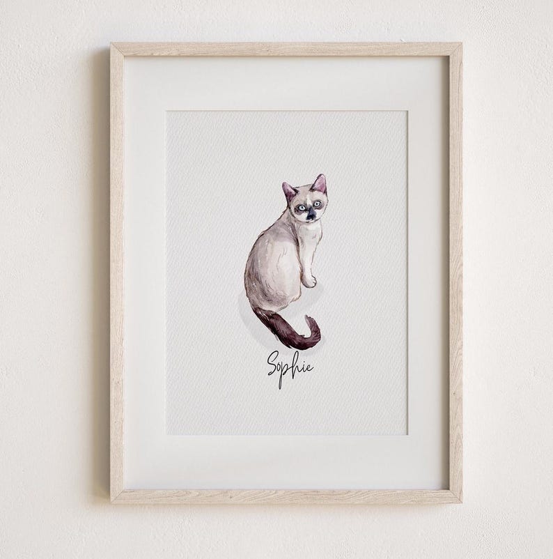 May include: Framed watercolour painting of a Siamese cat named Sophie. The cat is depicted in shades of grey, brown, and black, with blue eyes. The cat is sitting and looking towards the viewer. The name "Sophie" is written in cursive below the cat. The frame is light wood.