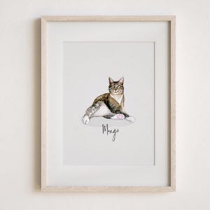May include: Watercolour painting of a cat in a light wood frame. The cat is brown, white, and grey, with the name "Mango" written below in cursive. The cat is lying down with its paws outstretched.