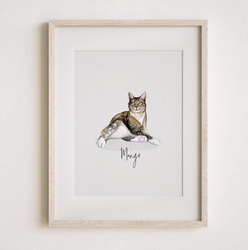 May include: Watercolour painting of a cat in a light wood frame. The cat is brown, white, and grey, with the name "Mango" written below in cursive. The cat is lying down with its paws outstretched.